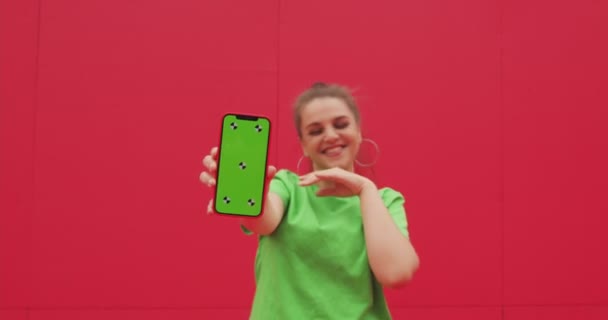 Muchacha joven feliz sonriente moderna mostrando el teléfono con la pantalla verde de la llave del croma con los puntos de seguimiento, divirtiéndose por la pared roja — Vídeos de Stock