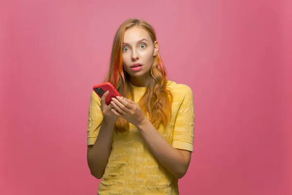 Sorprendida joven sorprendida con el teléfono asombrado por el puesto de noticias en la red social en el fondo de estudio rosa — Foto de Stock