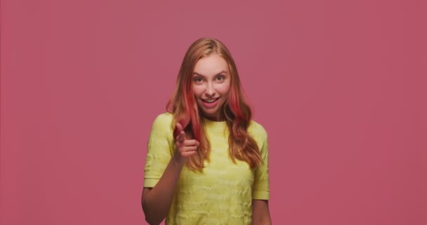 Усміхнена молода дівчина вказує вам і фотоапарат з пальцями, вибираючи щасливий, показуючи напрямок на рожевому фоні — стокове відео