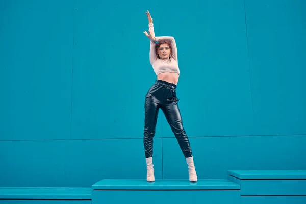 Модная девушка, стоящая с поднятой рукой на подиуме у синей стены с копировальным местом для рекламы. Сексуальная модель женщины позирует на открытом воздухе — стоковое фото