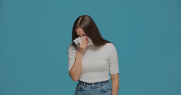 Menina insalubre espirrando em guardanapo, sentindo-se mal, sofrendo de sintomas de gripe ou alergia temporada, fundo estúdio azul — Vídeo de Stock