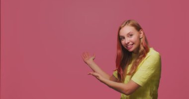 Reklam için boş alan gösteren genç bir kız pembe stüdyo arka planında hoş geldin jesti yapıyor.