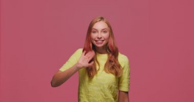 Gülümseyen genç kız el sallayarak merhaba ya da güle güle diyor. Sosyal ve mutlu kadın karşılaması, pembe arka planda selamlaşma.