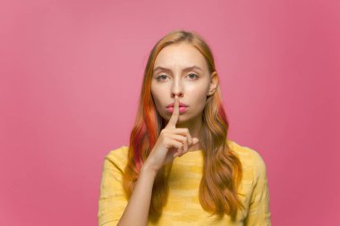 Güzel, ciddi, genç bir kız sessiz ve gizli kalmak istiyor. Pembe stüdyo arka planında sessiz bir jest yapıyor.