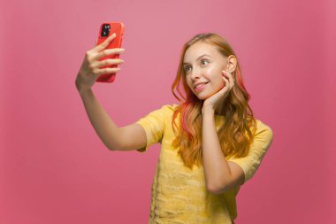 Modern, gülümseyen mutlu kız cep telefonuyla özçekim yapıyor. Sosyal ağ içeriği için pembe stüdyo arka planında.