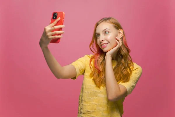Moderno joven sonriente feliz chica haciendo selfie disparo en el teléfono móvil para el contenido de la red social en rosa fondo del estudio — Foto de Stock