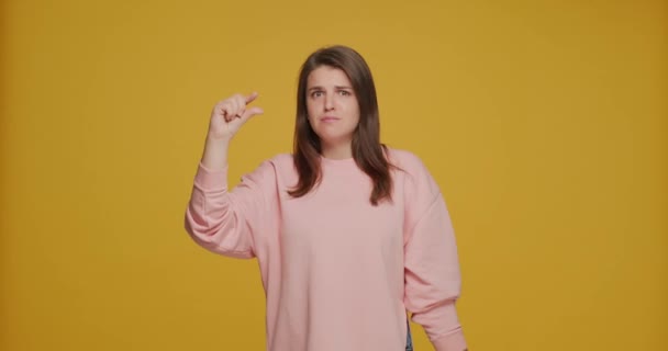 Vicces fiatal lány bemutató kis méret vagy kis gesztus ujjaival, látszó könyörgő sárga stúdió háttér — Stock videók
