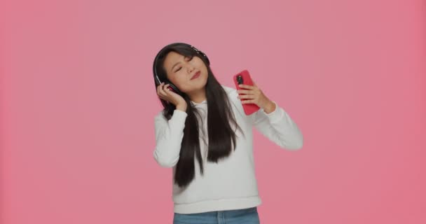 Asiatisches Mädchen hört per Kopfhörer Musik und hat Spaß beim Tanzen mit mobilen Musik-Apps auf rosa Hintergrund — Stockvideo