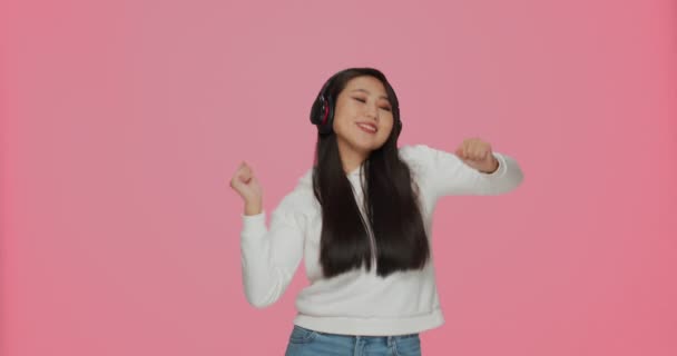 Dansa ung asiatisk flicka i hörlurar njuta lyssna nya coola modern musik mix på rosa studio bakgrund — Stockvideo