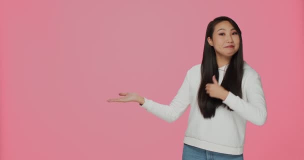 Usmívající se krásná asijská dívka ukazuje reklamní prostor s dobrou prodejní nabídku nebo se zabývají na růžové pozadí studia — Stock video