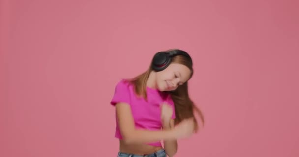 Cute little kid girl listen to music with wireless headphones, dancing, having fun on pink studio background — Stock Video