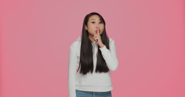 Pretty asian young girl showing silence gesture keep finger at lips ask be quiet, make secret offer on pink background — Stock Video