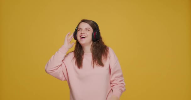 Moderna chica joven feliz escuchando música con auriculares, bailando, divirtiéndose en el fondo del estudio amarillo — Vídeos de Stock