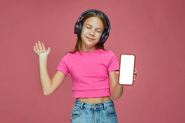 Iklan aplikasi mobile musik. Happy little girl child in headphone hold phone with white empty screen mock up — Stok Foto