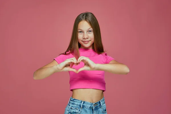 Happy cute gadis kecil membuat tanda hati dengan tangan, menunjukkan sikap cinta, mengungkapkan perhatian pada latar belakang merah muda — Stok Foto