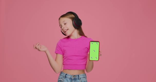Музыкальные мобильные приложения. Симпатичная маленькая девочка в наушниках любит слушать музыку, держа в руках телефон с зеленым экраном — стоковое видео