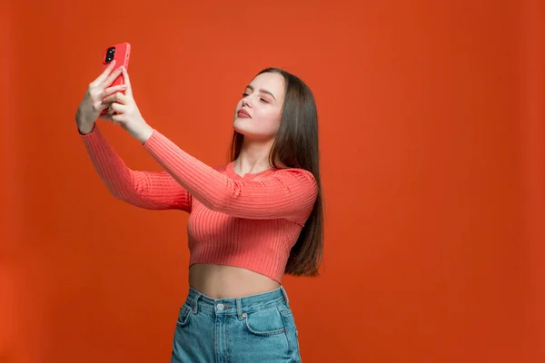 Young girl making selfie on phone. Pretty woman making faces on camera, posing for picture on orange studio background — Stock Photo, Image