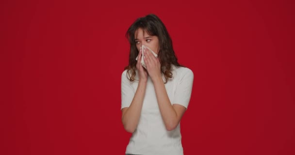 Нездоровая молодая девушка чихает, сморкаясь в салфетку, страдает бегущим носом, симптомами гриппа, сезонной простудой на красном фоне — стоковое видео