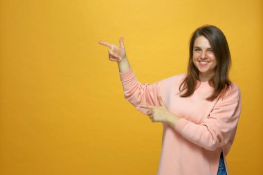 Gülümseyen genç kız sarı arka planda telif alanını işaret ediyor, reklam yapıyor, online mağazada ürün öneriyor.