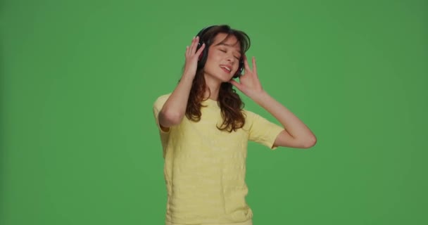 Güzel genç kız kulaklıkla müzik dinliyor, sesten hoşlanıyor, yeşil renkli stüdyo arka planında dans ediyor. — Stok video