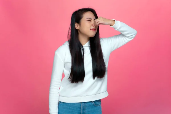 Bad smell. Young asian girl holding breath with fingers, pinch nose from stinky odor on pink studio background
