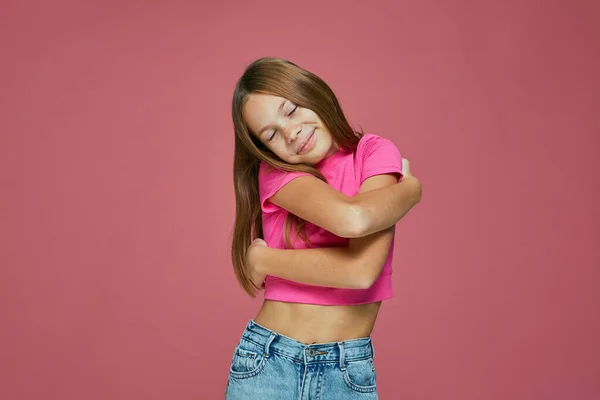 快乐的小女孩依偎在粉红工作室的背景下,用爱心和关怀拥抱自己.健康儿童的自尊概念 — 图库照片