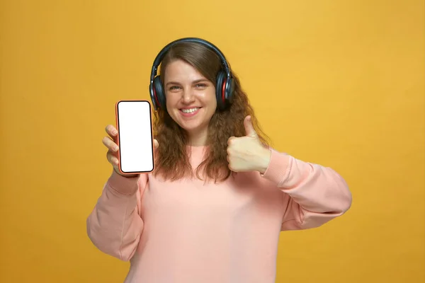 Moderne jeune fille heureuse dans les écouteurs montrent téléphone avec écran blanc maquette, recommander l'application mobile de musique, ne pouce vers le haut — Photo