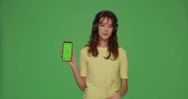 Kulaklıklı mutlu genç kız yeşil ekranlı telefon gösteriyor, krom arka planda müzik mobil uygulamasının keyfini çıkarıyor.