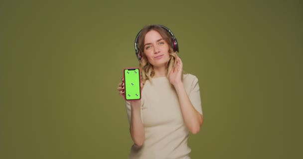 Счастливая женщина в наушниках показывает телефон с макетом зеленого экрана. Девочки наслаждаются музыкой, танцами, мобильным приложением — стоковое видео