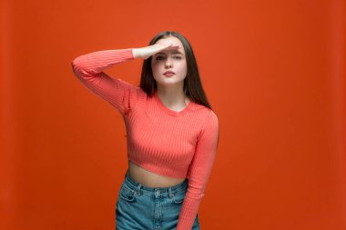 Dikkatli meraklı genç kız, elini alnına uzatıp uzaklara bakıyor. Turuncu arka planda yeni şeyler keşfediyor.