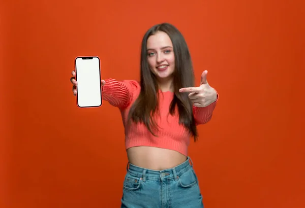 Elegante chica feliz apuntando en el teléfono con la pantalla vacía maqueta sobre fondo naranja. Compras en línea, compra, nueva aplicación — Foto de Stock