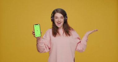 Kulaklıklı gülümseyen genç kız yeşil ekran ile telefon gösteriyor, müzik dinliyor. Müzik uygulamaları reklamı 