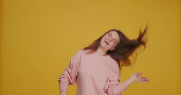 Enerjik mutlu genç kız saçlarını sallıyor, gülüyor, dans ediyor, sarı stüdyo arka planında eğleniyor. — Stok video