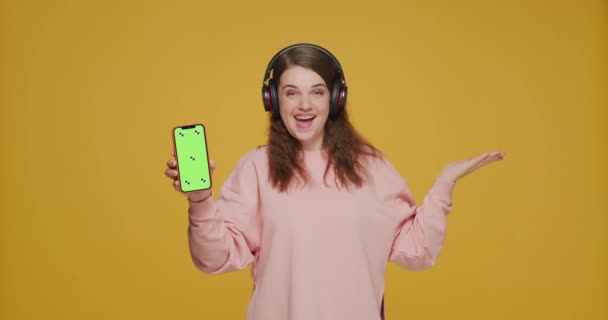 Улыбающаяся молодая девушка в наушниках показывает телефон с макетом зеленого экрана, слушает музыку. Реклама музыкальных приложений — стоковое видео