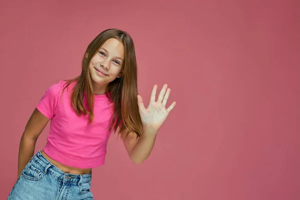 Freundliche Kinderbloggerin winkt mit der Hand in Grußgeste, lächelnd begrüßt Abonnenten auf rosa Studiohintergrund — Stockfoto