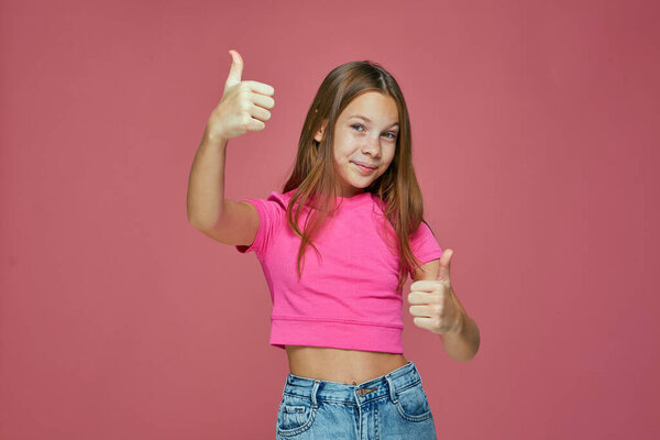 Счастливая девочка показывает большой палец вверх, рекомендует услугу или продукт, просит поставить лайки на розовом студийном фоне