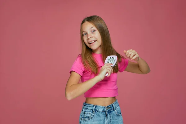 Beautiful child girl combing long hair make coiffure hairstyle, preening on pink studio background. Children haircare — 图库照片