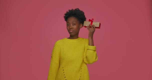 Όμορφη νεαρή Αφρικάνα Αμερικάνα κουνούσε το κουτί με τα δώρα, κρατώντας το αυτί της κοντά και μάντεψε τι μέσα, χαμογελώντας — Αρχείο Βίντεο