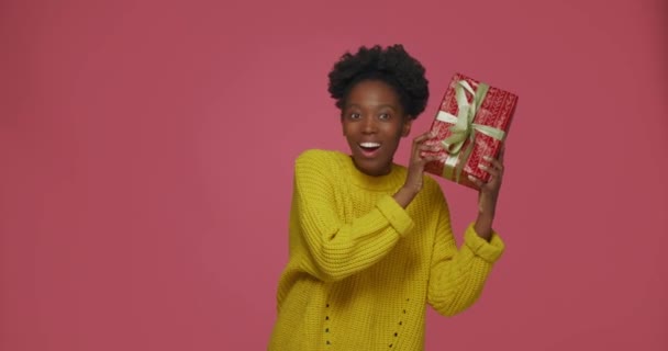 Happy friendly african american young girl shaking wrapped gift box, holding near ear listening to guess what inside — Stock Video