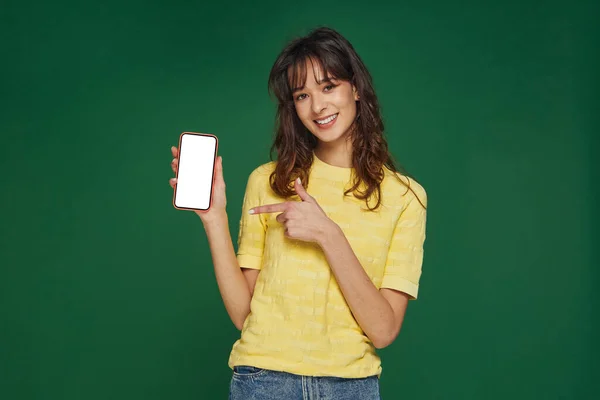 Chica joven y feliz señalando con el dedo en el teléfono de pantalla en blanco. Chica recomiendan aplicación moderna tienda móvil en línea — Foto de Stock