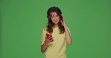 Müzikal mobil uygulamalar reklamı. Kulaklık takan genç modern kız, müzik dinlemeyi seviyor. Kromakey yeşil arka planda telefon kullanarak.