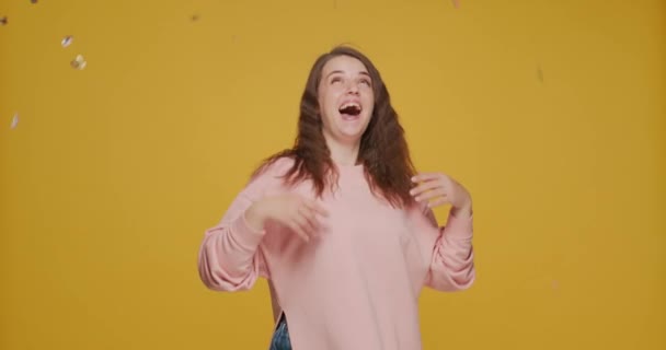 Chica joven feliz divirtiéndose bajo confeti, celebrar las vacaciones en el fondo del estudio amarillo. Concepto de venta festiva — Vídeos de Stock