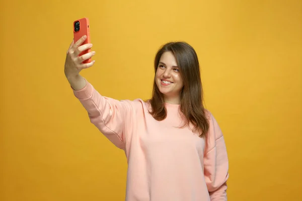 Happy girl taking selfie holding phone looking at smartphone using mobile camera smiling having fun on yellow background — Stock Photo, Image