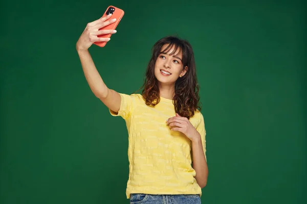 Beautiful young girl blogger posing, taking selfie photo on phone for social network content on green studio background — Stock Photo, Image