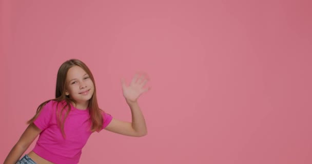 Sonriente niña saludo, saludar con la mano saludar o adiós a los suscriptores, echar un vistazo en el fondo rosa con espacio de copia — Vídeos de Stock