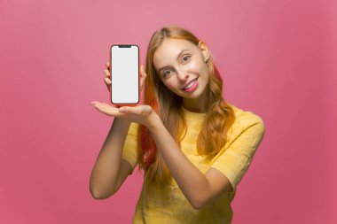 Gülümseyen genç bir kadın pembe stüdyo arka planında boş bir telefon ekranı gösteriyor. Modern mobil uygulamalar reklamcılığı