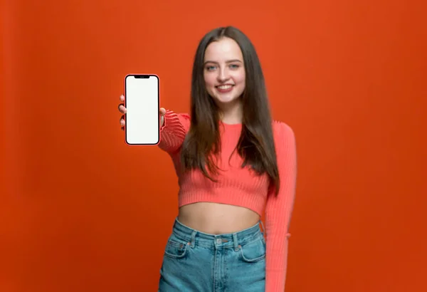 Sorrindo menina moderna mostrar telefone inteligente com tela de simulação em branco no fundo laranja. Aplicativos móveis publicidade — Fotografia de Stock