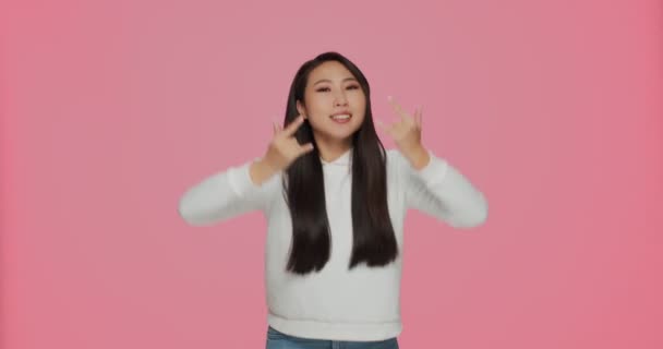 Крутая азиатская молодая девушка рокер показывая рок-н-ролл рога руки жест с веселой палкой языка на розовом фоне студии — стоковое видео