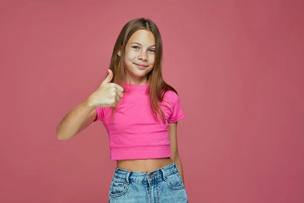 Pozitív gyermek lány mutató hüvelykujj fel gesztus, mint a jel, mosolygós ajánlom jó vagy szolgáltatás rózsaszín stúdió háttér — Stock Fotó