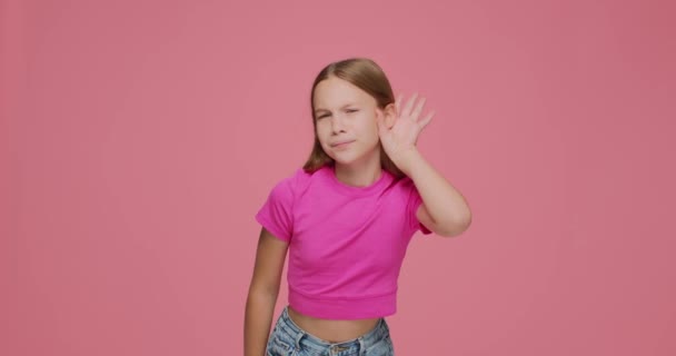 Спантеличена дівчинка кладе руки на вуха, щоб слухати краще, сміючись над анекдотом на рожевому фоні. Проблеми зі слухом — стокове відео